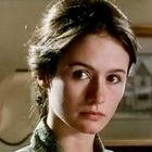 Eine junge Frau mit ernstem Ausdruck, im Hintergrund ein unscharfes Bild eines Hauses.