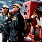 Zwei Männer in Winterjacken mit "U.S. Forest Service"-Abzeichen, ein Hund in einem roten Fahrzeug, schneebedeckte Landschaft.