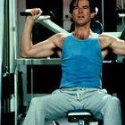 Eine Person im blauen Tanktop trainiert an einer Schulterpresse im Fitnessstudio.