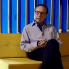 Eine Person sitzt auf einem gelben Sofa vor einem Hintergrund aus vertikalen blauen Streifen. Sie trägt eine Brille und Halstuch.