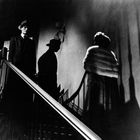 Zwei Schattenfiguren stehen auf einer Treppe, eine im Anzug und Hut, die andere in einem Mantel, vor einem dunklen Hintergrund.