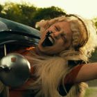Person mit blondem Haar und dunklem Lippenstift lehnt sich lachend aus einem Autofenster. Hintergrund zeigt Natur.