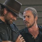 Zwei bärtige Männer in Westernkleidung, einer mit Hut. Der linke Mann lächelt, der rechte scheint zu sprechen.