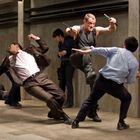 Ein Mann in einem schwarzen Tanktop kämpft mit zwei Männern in Anzügen in einem betonierten Raum. Dynamische Kampfszene.