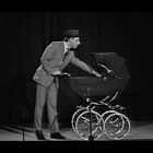 Ein Mann in Anzug und Hut greift in einen klassischen Kinderwagen mit großen Rädern, alles in Schwarz-Weiß gehalten.