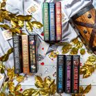 Sieben Bücher von Jennifer Lynn Barnes liegen auf einem satinierten Stoff, umgeben von goldenen Blättern und Spielkarten.