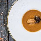Ein Teller mit cremiger Suppe, dekoriert mit einem Stück Brot und frischen Sprossen; daneben rote Linsen auf Holz.