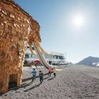 Holz-Mammut mit Rutsche, Kinder laufen darunter. Moderne runde Hütte, Berge und strahlender Sonnenschein im Hintergrund.