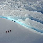 Drei Personen wandern in leuchtender Ausrüstung über eine verschneite Eisfläche, unterhalb beeindruckender Gletscherstrukturen.