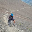 Eine Person mit blauem Helm schiebt ein Fahrrad einen steilen, steinigen Bergpfad hinauf. Der Pfad schlängelt sich nach oben.