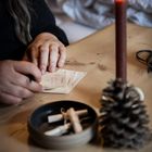Mehrere beschriftete Samenpäckchen, eine Person schreibt. Eine dunkle Kerze in einer Tannenzapfenhalterung brennt.