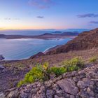Die Ansicht zeigt eine ruhige Küste bei Sonnenuntergang, mit blauem Meer, felsiger Landschaft und vereinzeltem Grün in der Nähe.