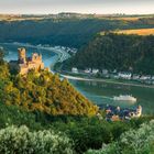 Ein Fluss windet sich durch ein grünes Tal. Eine imposante Burg steht links, im Hintergrund Hügel und Dörfer.