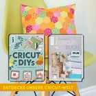 Zwei Bücher über Cricut-Projekte vor einem bunten Kissen auf einem gelben Stuhl, darunter steht: "ENTDECKE UNSERE CRICUT-WELT".