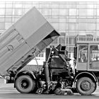 Ein Reinigungsfahrzeug mit angehobenem Container zeigt seine Kehrbürsten, im Hintergrund ein Gebäude aus Glas.
