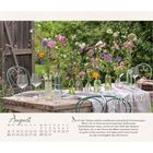 Augustkalender mit Blütenvasen auf einem Holztisch im Garten, umgeben von grünen Stühlen und Gläsern.