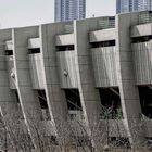 Großes Betongebäude mit den olympischen Ringen. Im Vordergrund sind kahle Bäume. Hochhäuser im Hintergrund.