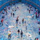 Menschen bemalen eine große, ovale Fläche und Wände mit bunten Farben. Es herrscht reges Treiben und kreative Aktivität.