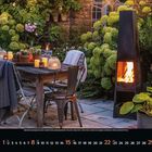 August 2023.  

Ein einladender Garten mit Tisch und Stühlen, umgeben von grünen Hortensien und einem modernen Kamin mit Flammen.