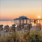 Strandkörbe am Sandstrand, Steg führt zu einem Gebäude im Meer, orange Sonnenuntergang am Horizont.
