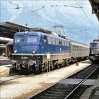 110 183-1 steht auf einer blauen Lokomotive am Bahnhofsgleis. Dahinter sind Berge und eine zweite Lokomotive zu sehen.