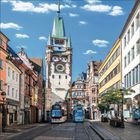 Eine Altstadtstraße mit bunten Gebäuden; im Zentrum ein historischer Turm mit einer großen Uhr und zwei Straßenbahnen.