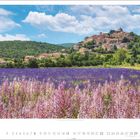 Text: "8" unten links. Kalender mit violettem Blütenfeld, dahinter ein Dorf auf einem Hügel, blauer Himmel mit Wolken.