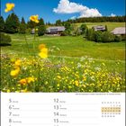 April 2027 Kalenderseite, blühende Wiese, gelbe und weiße Blumen, im Hintergrund Wälder und Gebäude unter blauem Himmel.