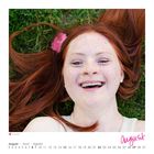 Kalender: August. Frau mit roten Haaren und Sommersprossen, lachend im Gras liegend.