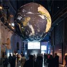 Texte: "BROWN BLUFF", "PARADISE", "KONTINENT CONTINENT". 
Große Erdkugel hängt im Raum, Besucher betrachten Ausstellungsstücke.