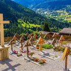Eine Ansammlung von Holzkreuzen auf einem Friedhof, umgeben von blühenden Blumen, vor einer grünen Berglandschaft.