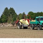 „August“, „Commander“, „Unimog 427 U 1600 Baujahr 2002“. Grün-gelbes Fahrzeug mit landwirtschaftlichem Anhänger auf einem Feld.