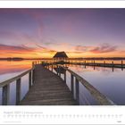 August 2027 Kalenderblatt, Schleswig-Holstein. Ein Holzsteg führt zu einem Pavillon über einem ruhigen See bei Sonnenuntergang.
