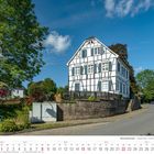 Kalenderblatt August 2027 mit Fachwerkhaus auf Anhöhe, umgeben von Bäumen und blühenden Blumen, klarer Himmel im Hintergrund.