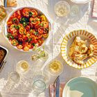 Tomatensalat, Brot, gebackenes Fleisch, Gläser mit Getränk auf einem Tisch mit weißem Tuch. Sonnenlicht fällt darauf.