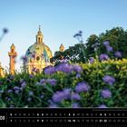 Wien Kalender für August: Die Karlskirche hinter lila Blumen und Bäumen bei blauem Himmel.