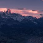 Bergkette bei Sonnenuntergang, Wolken mit rosa und violetten Tönen, darunter eine kleine Stadt im Tal.