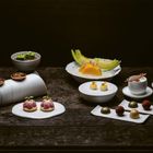 Eine elegante Dessertpräsentation mit Törtchen, Pralinen, Tee und Melonenstücken auf weißen Geschirrstücken.