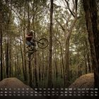 Kalendertage und Wochentage sind unten dargestellt. Ein Mountainbiker springt über eine Erdhügel in einem bewaldeten Gebiet.