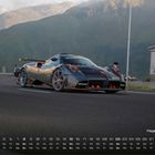 "Pagani Huayra 8" und Monatskalender. Sportwagen auf Bergstraße, Berge im Hintergrund, Gebäude rechts.