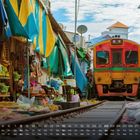 Ein Kalender zeigt einen farbigen Zug, der durch einen engen Markt mit bunten Planen fährt. Menschen stehen dicht dabei.