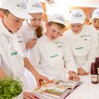 Kinder in Kochuniformen mit "Europa Miniköche" Mützen schauen in ein Kochbuch. Daneben Pflanzen und Flaschen.