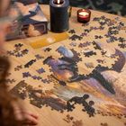 Ein Tisch mit einem teilweise zusammengesetzten Puzzle zeigt Drachen. Kerzen brennen daneben. Eine Person ist anwesend.