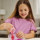 Ein Kind in einem rosa T-Shirt gießt rosa Flüssigkeit aus einem Becher in eine transparente Form auf einem Tisch.