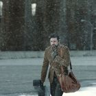 Keine lesbaren Texte. Ein Mann geht durch Schnee, trägt einen Mantel und hält eine Gitarrentasche und braune Ledertasche.