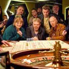 Personengruppe in einem Casino steht gespannt um einen Roulettetisch. Das Rad dreht sich, und alle beobachten konzentriert.