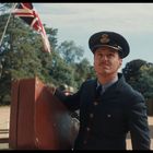 Zwei Männer in Militäruniformen; einer trägt eine große Tasche. Im Hintergrund wehen britische Flaggen vor Bäumen.