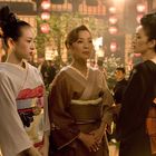 Ich kann den Text nicht lesen. Drei Frauen in kimonos stehen in einem traditionell japanischen Raum mit roten Laternen.