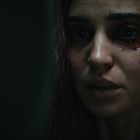 Eine Frau mit blutigem Auge und ernstem Ausdruck im dunklen, düsteren Licht.