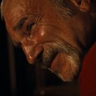 Ein älterer Mann mit grauem Haar und Schnurrbart lächelt in einem dunklen Raum. Er trägt ein rotes Oberteil.